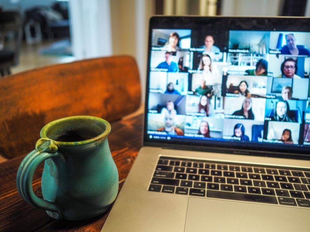 A laptop sitting on a wooden table, with a virtual meeting on the screen. There are a lot of attendees with their cameras on, and there is a coffee mug next to the laptop.